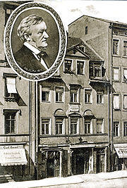 A postcard of a five-story building with shops on the ground floor and garret windows in the roof. A round inset has a picture of Wagner in middle age.