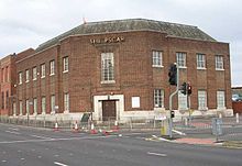 A plain corner building of the 1930s on a main road. Traffic-lights, railings, bollards and pedestrian crossings with tactile paving can also be seen. The frontage of the building is mainly brick, but the lowest course, of Portland stone, extends as far as the sills of the eleven ground-floor windows (eight on the main road and three on the side road to the right). The wooden double entrance door set diagonally on the corner also has a stone surround. Above it is a long window with a projecting brick surround, and above this is a large sign in capital letters, originally reading SHEEPSCAR but now with the second