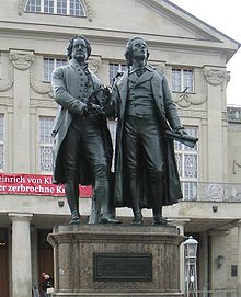 Photograph of a large bronze statue of two men standing side-by-side and facing forward. The statue is on a stone pedestal, which has a plaque that reads